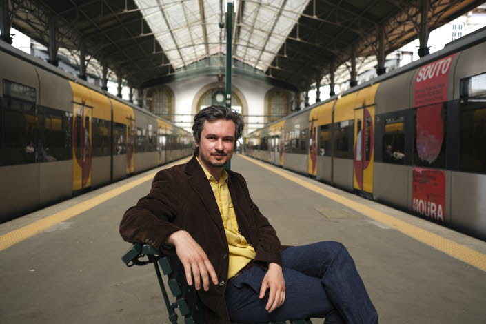 Bild på Maxim på en tågperrong med två tåg bredvid. Han sitter på en stol med gul skjorta och mörk kavaj. 