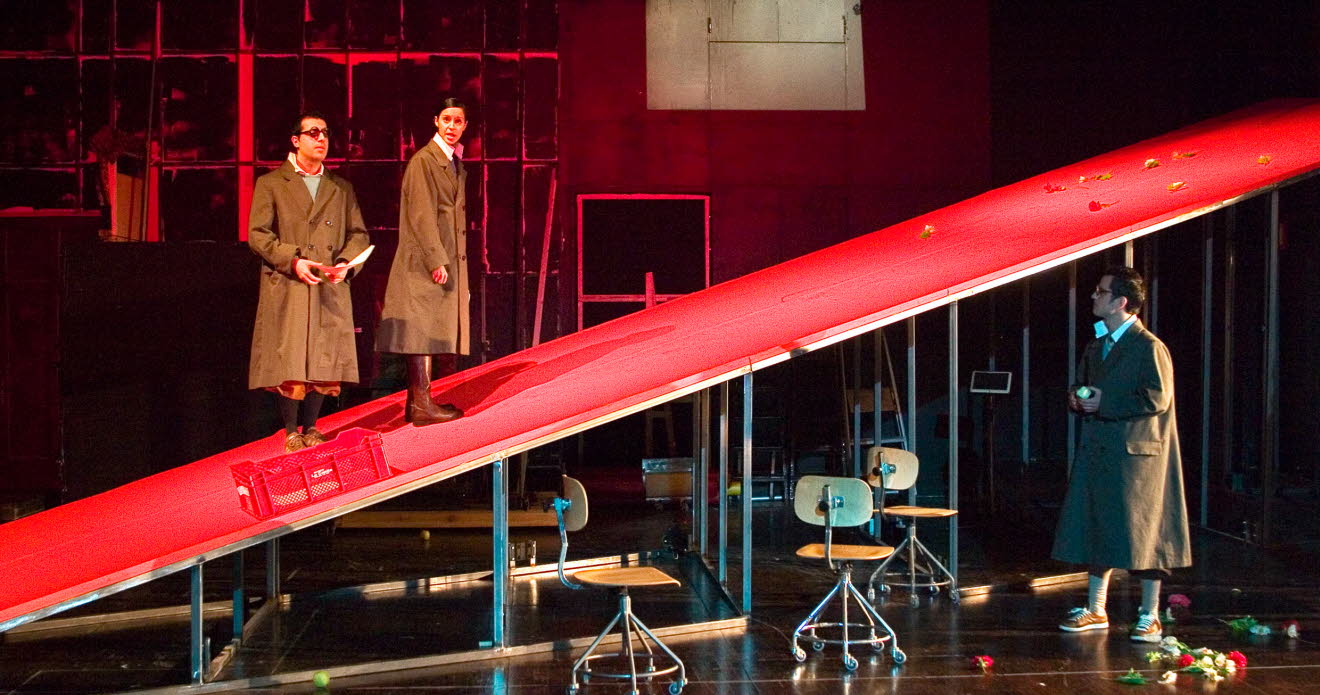 A theatre scene, one man and one woman are standing on a red ramp, another man looks up at them. 