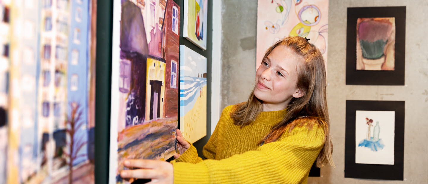 Ett barn rättar till en tavla i ett rum fyllt av målningar. Hon ser glad ut.