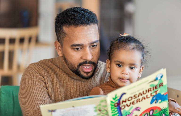 En liten flicka sitter i en mans knä som pekar och läser ur en bok om dinosaurier. Flickan tittar koncentrerat i boken. 