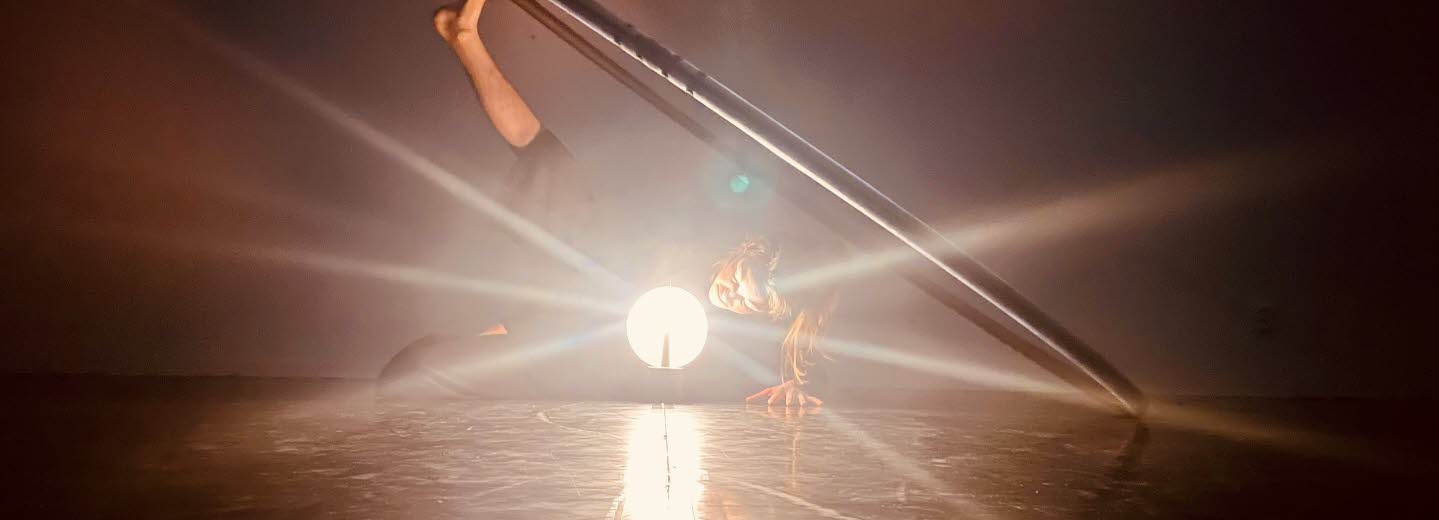 En kvinnlig dansare ligger på sidan i ett mörkt rum. Det ena benet är uppsträckt där foten balanserar en stor ring. Framför henne står en lampa som likt en strålkastare riktas mot betraktaren.