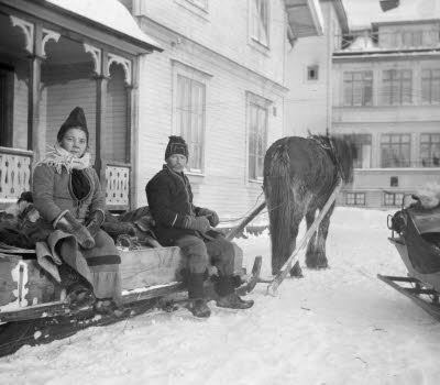 Svartvit bild, en häst drar två personen i en stor släde, framför ett gammalt hus. Det snöar lätt. 