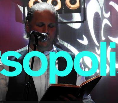 Turkos text med Versopolis, i bakgrunden en man som läser ur en bok på scen..