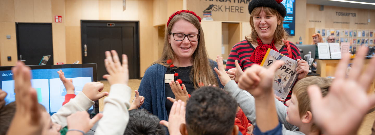 Två kvinnor står vända mot kameran i biblioteksmiljö. Framför dem, med ryggen mot kameran räcker många små barn upp händerna.