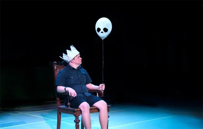 En man sitter på en scen. På huvudet har han en kungakrona i papp, i handen håller han en ballong med spökansikte.