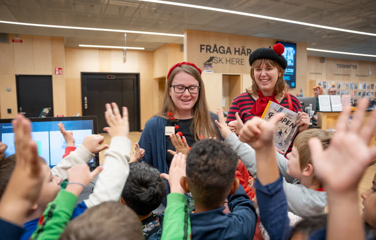 Två kvinnor står vända mot kameran i biblioteksmiljö. Framför dem, med ryggen mot kameran räcker många små barn upp händerna.
