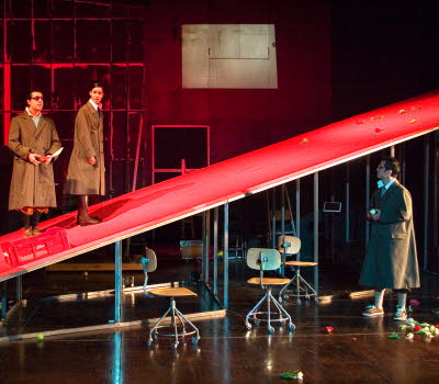 A theatre scene, one man and one woman are standing on a red ramp, another man looks up at them. 