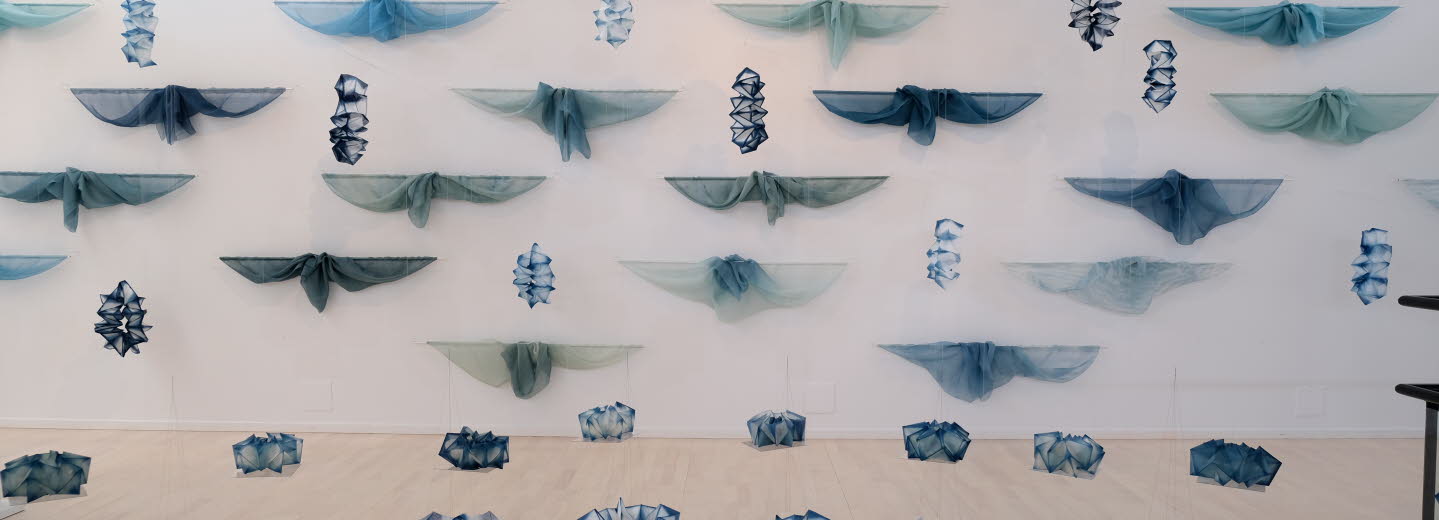 Konstinstallation med blå och gröna tygstycken upphängda i rader på en vit vägg, samt geometriska former i liknande färger placerade på golvet.