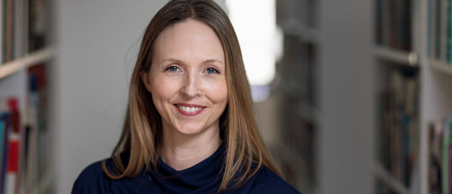 Medelålders kvinna med långt cendréfärgat hår och blå tröja omgiven av bokhyllor.