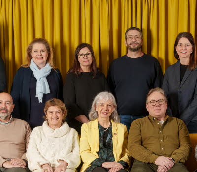 Gruppbild på jurymedlemmar, fem personer sitter i en gul soffa, bakom soffan står sex personer, i bakgrunden hänger en stor gul gardin. 