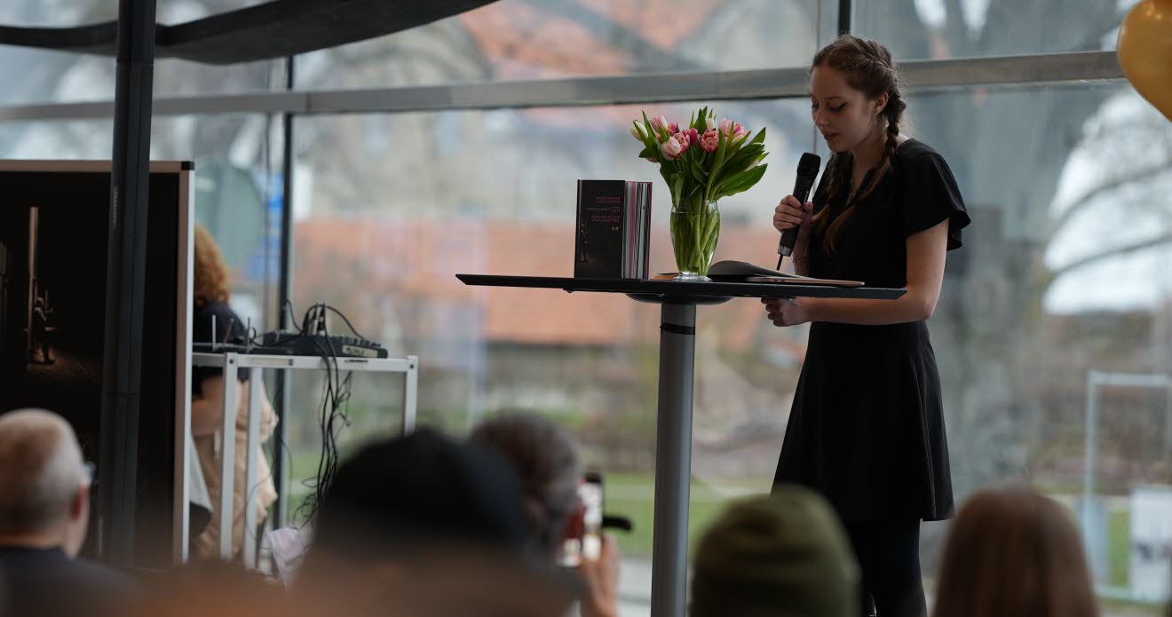 En ung kvinna står med en mikrofon på en scen och läser ur en bok.