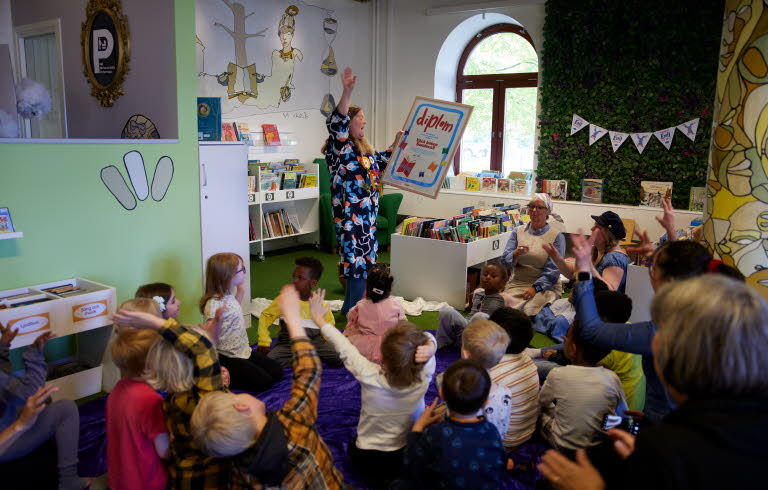Barn som sitter i en halvcirkel kring en kvinna som håller upp ett stort diplom. 