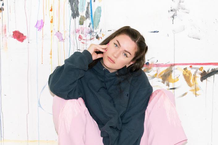 Artist Moa Romanova is sitting against a wall in pink pants and a black hoodie. 