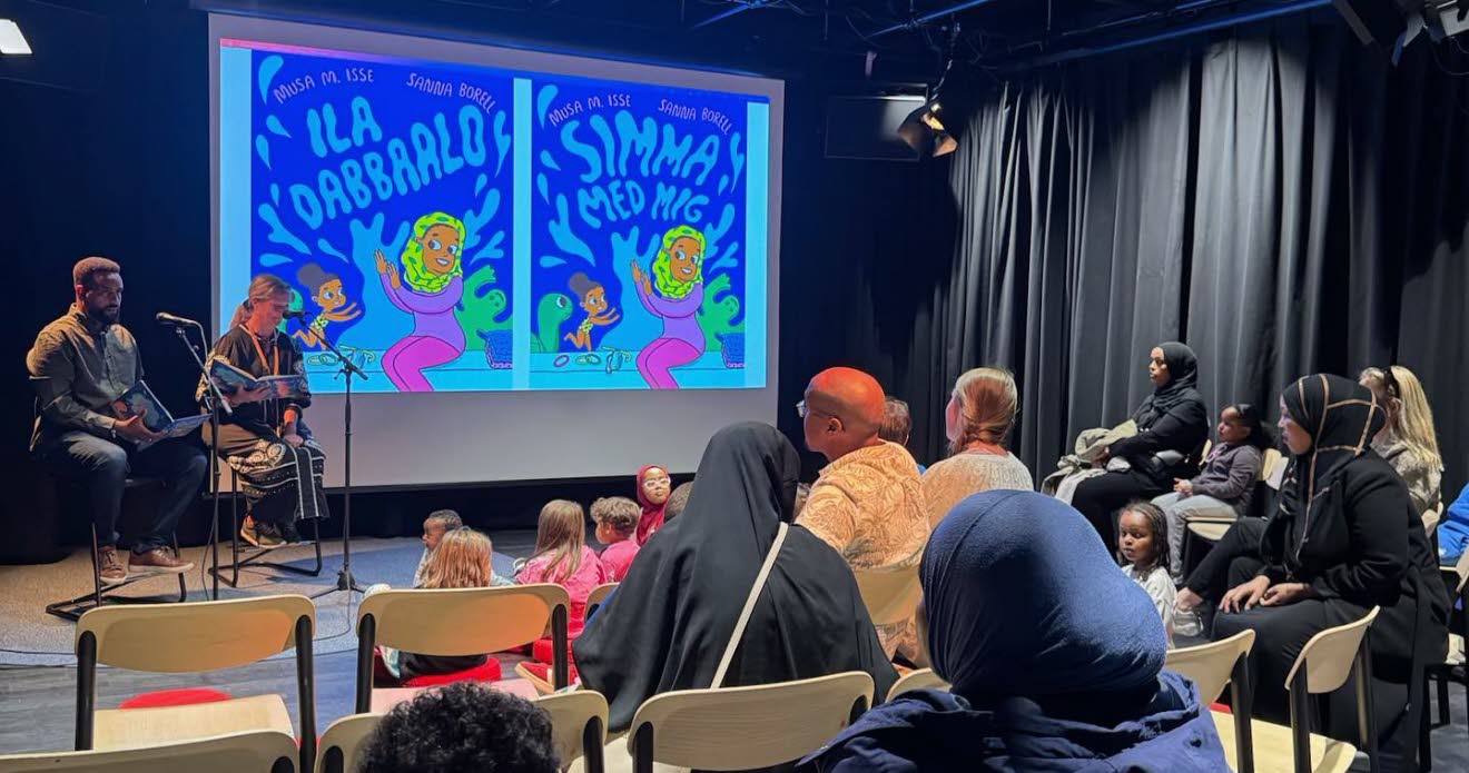 Två personer sitter på en scen och läser framför en publik med barn och vuxna.