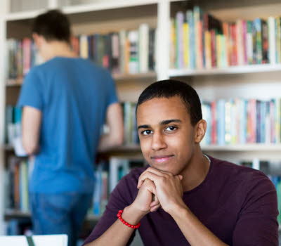 Tre ungdomar på bibliotek, en kille sitter vid ett skrivbord och studerar medan en tjej och en kille står vid en bokhylla och letar böcker. 