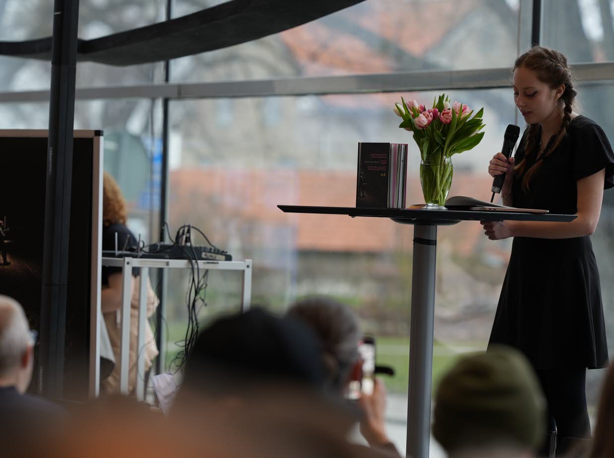 En ung kvinna står med en mikrofon på en scen och läser ur en bok.