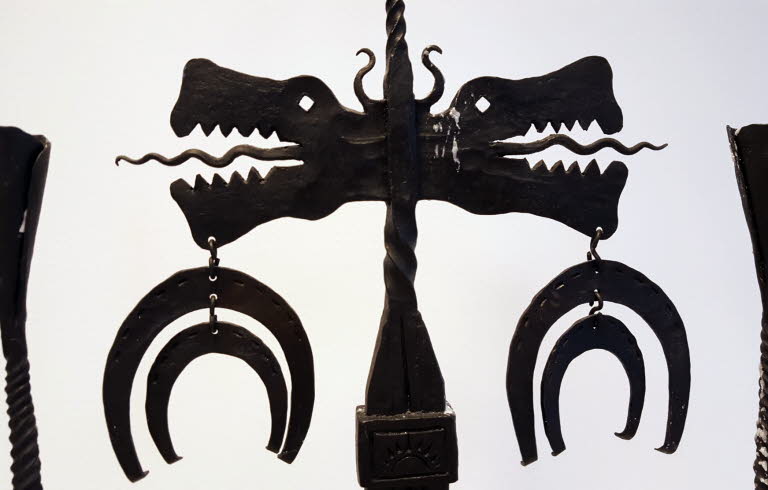 Överdelen av en ljusstake i smide med två drakhuvuden och hästskoformade hängen.