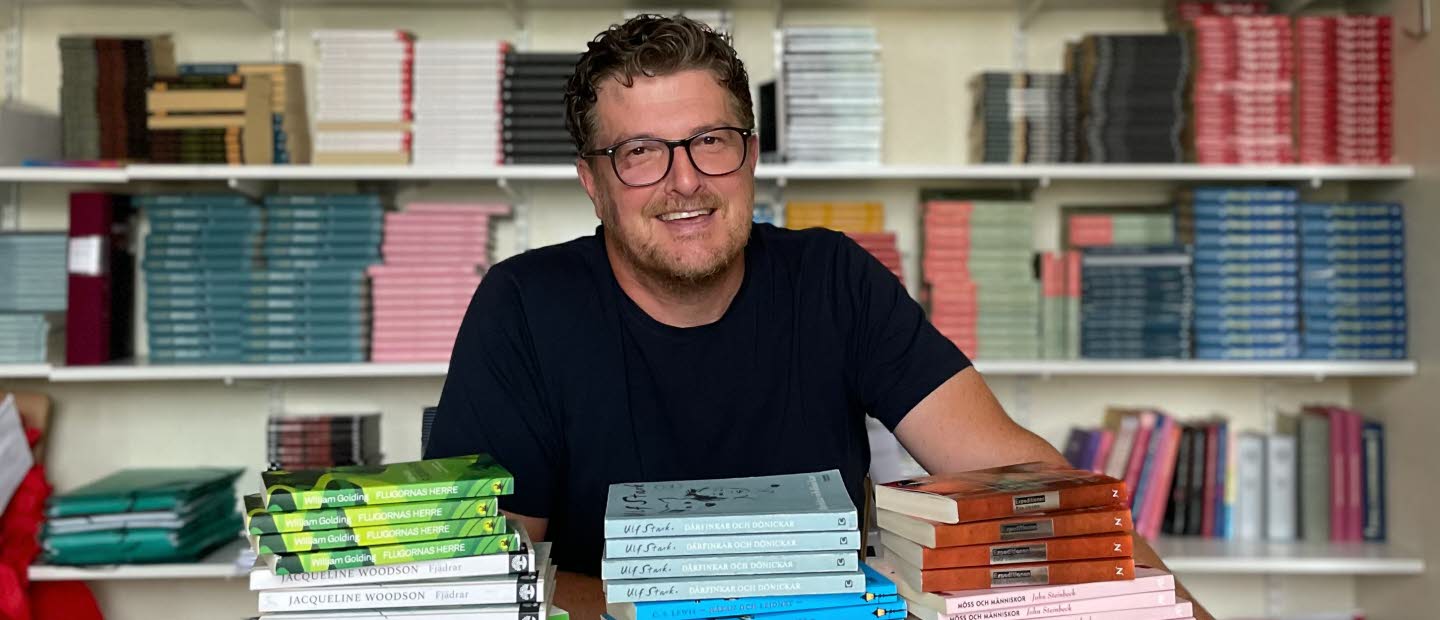 En leende man som sitter vid ett bord med travar av böcker. I bakgrunden bokhyllor fyllda med böcker. 
