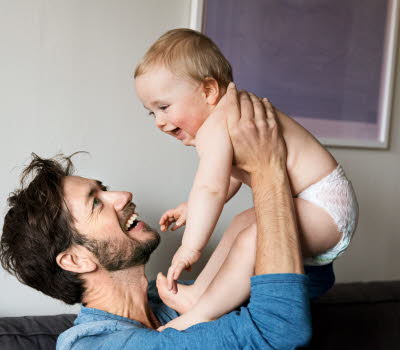En glad man håller upp en glad bebis i blöja. De ser ut att sitta i deras vardagsrum.