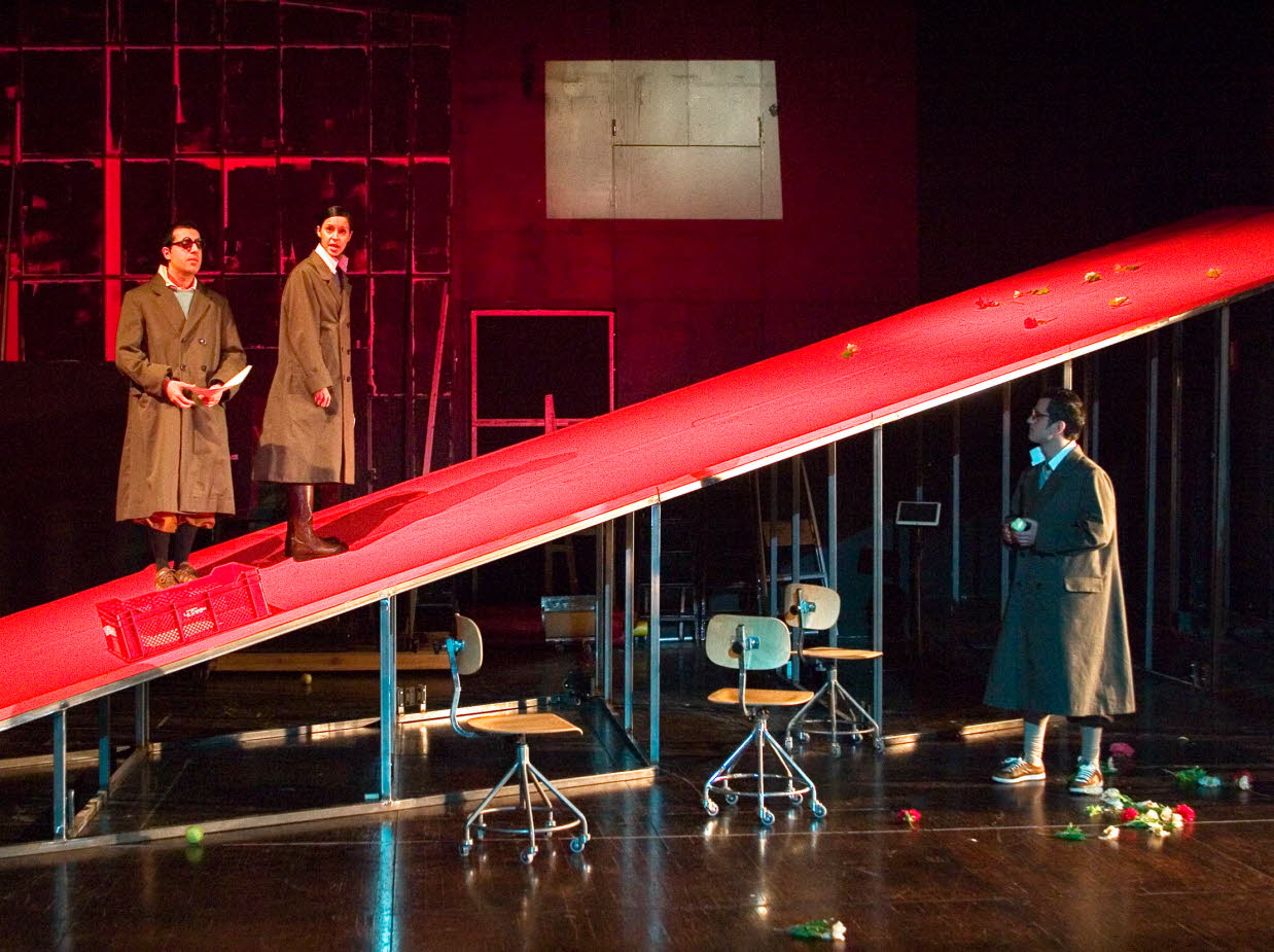 A theatre scene, one man and one woman are standing on a red ramp, another man looks up at them. 
