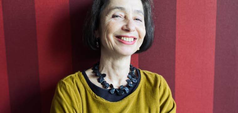 Foto på Elena Balzamo som ler mot kameran. Elena har en mörk page och är klädd i en senapsgul klänning med ett stort svart halsband. Bakgrunden är randig i röda toner. 