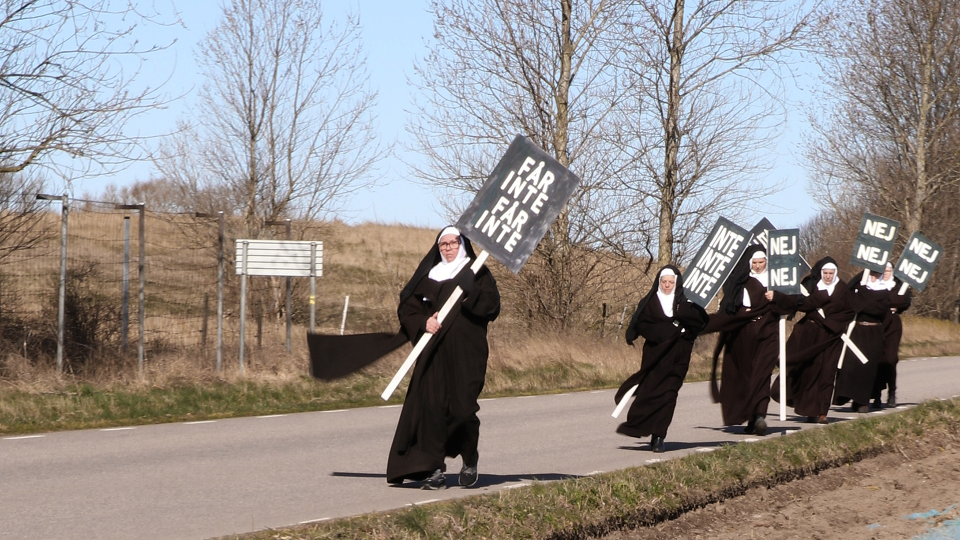 Nunnor går på en väg med plakat där det står "Får inte, får inte", "Nej, nej, nej". Det är höst eller tidig vår.