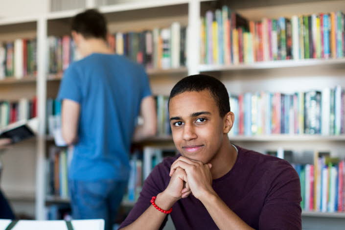 Tre ungdomar på bibliotek, en kille sitter vid ett skrivbord och studerar medan en tjej och en kille står vid en bokhylla och letar böcker. 