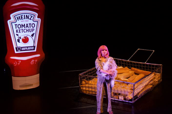 En tjej i rosa hår står på en mörk scen, i bakgrunden syns en förstorad ketchupflaska och förstorade pommes frites..