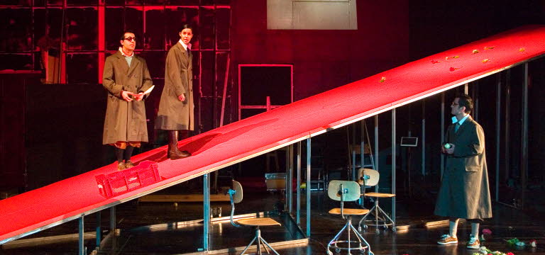 A theatre scene, one man and one woman are standing on a red ramp, another man looks up at them. 