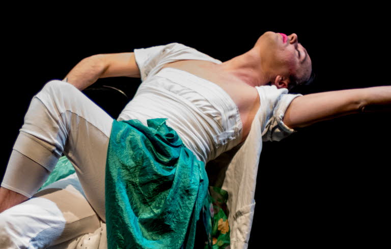 En dansare som sitter på en stol och gör en rörelse bakåt. Långa textiler som släpar på scengolvet. 