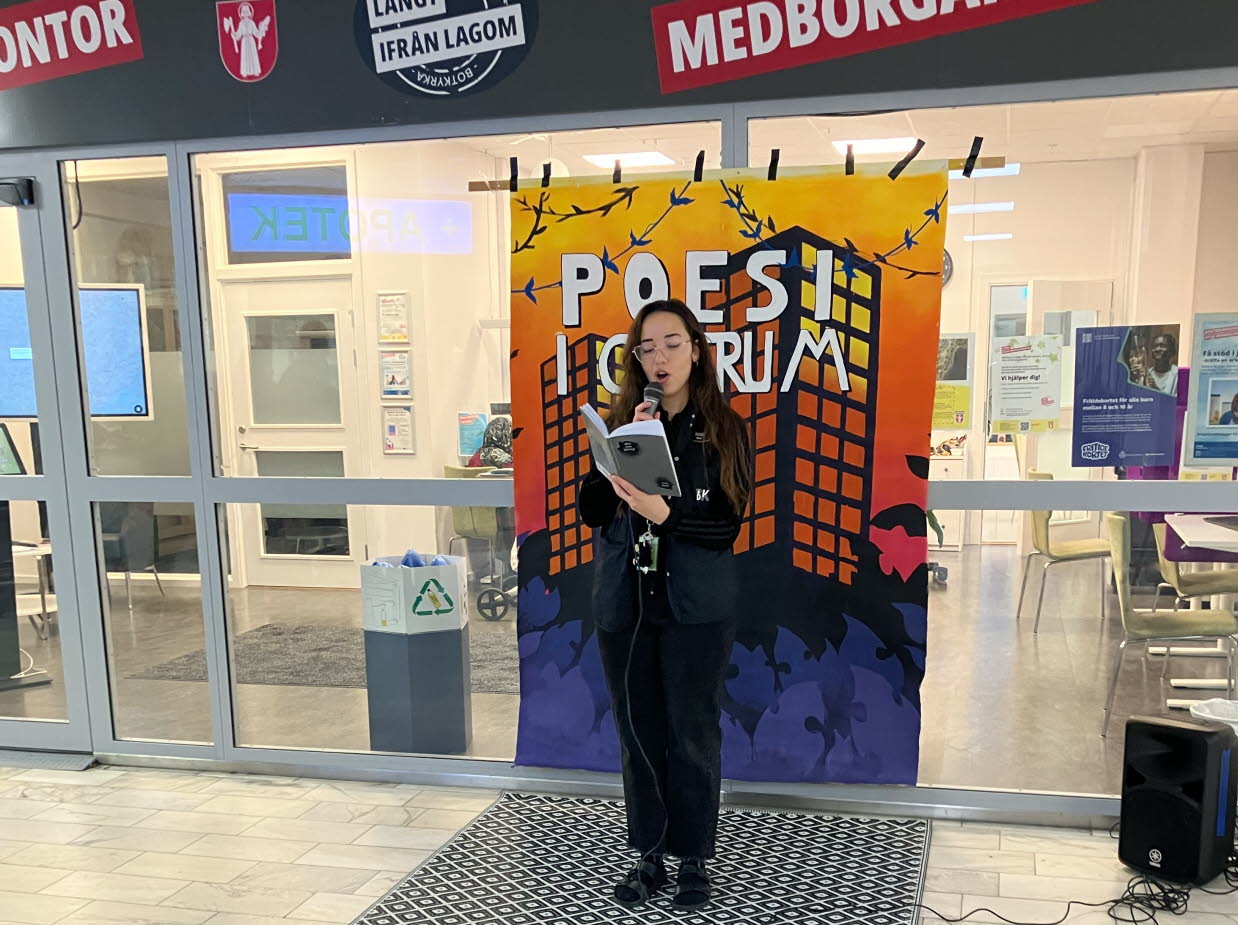 En ung kvinna står med en mikrofon i ett köpcentrum och läser ur en bok.