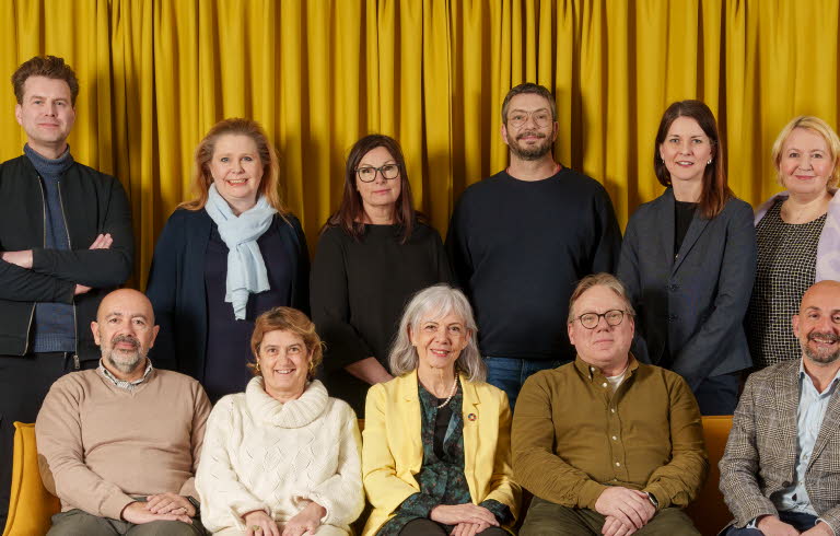 Gruppbild på jurymedlemmar, fem personer sitter i en gul soffa, bakom soffan står sex personer, i bakgrunden hänger en stor gul gardin.