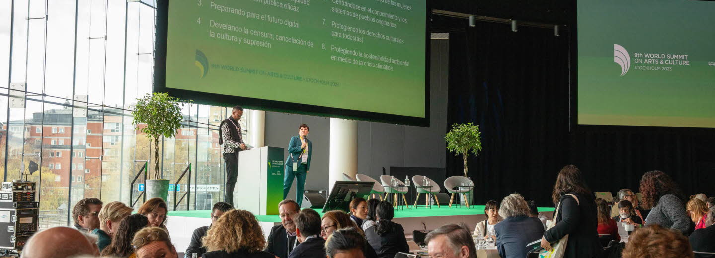 Vy över konferenslokalen och deltagare som sitter i grupper kring bord. På scenen två talare och en stor skärm som visar en presentation. 