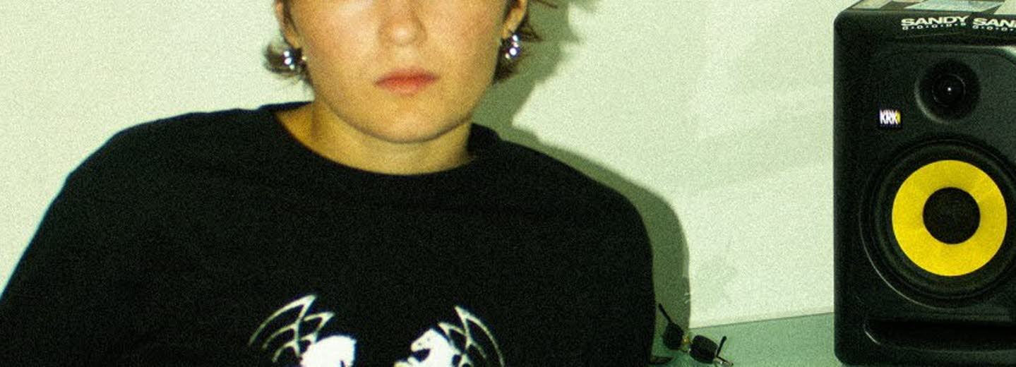 Kvinna i 20-årsåldern sittandes vid ett bord framför en laptop. På bordet en högtalare och hörlurar. 