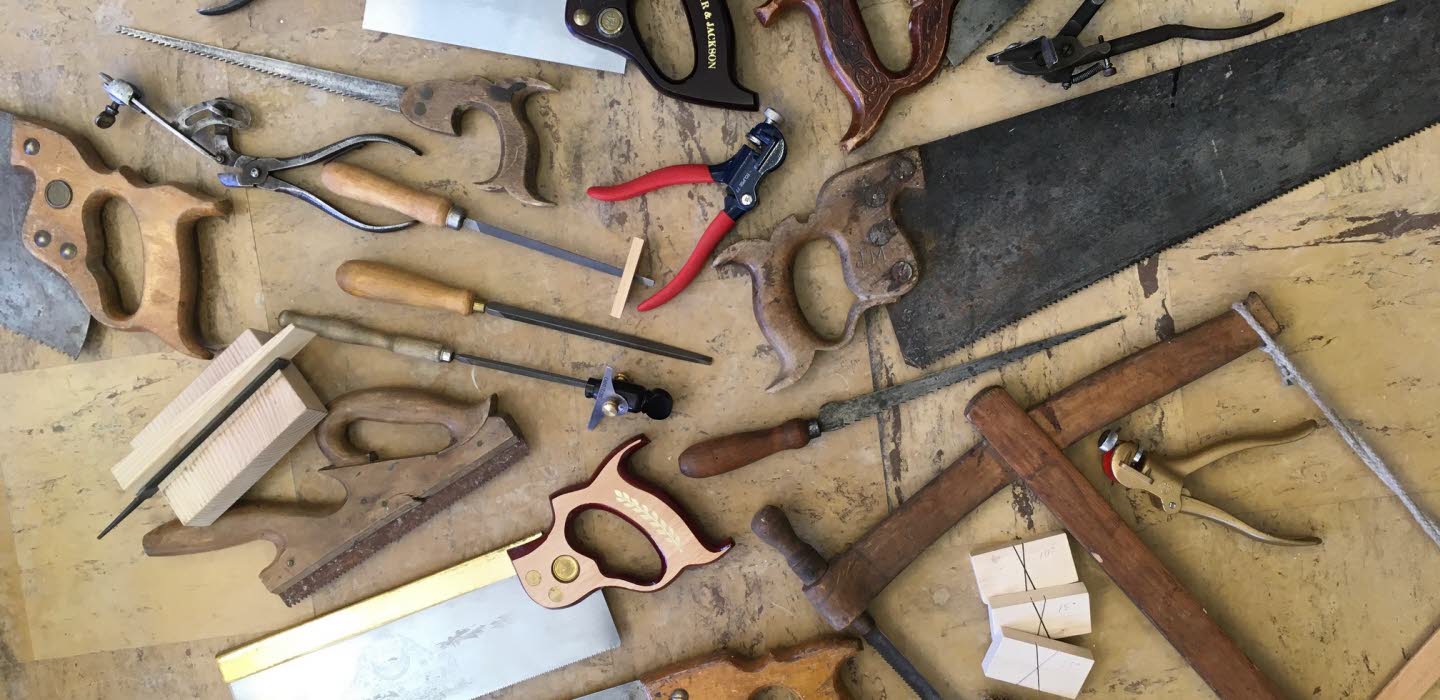 Gamla och nya handverktyg, främst sågar, filar och tänger, utspridda på ett slitet träbord.