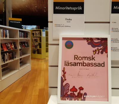 Intyg på att Umeå bibliotek har blivit Romsk läsambassad. Intyget står på ett bord, bokhyllor i bakgrunden. 