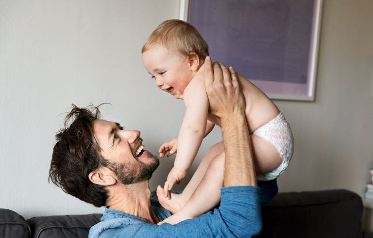 En glad man håller upp en glad bebis i blöja. De ser ut att sitta i deras vardagsrum.