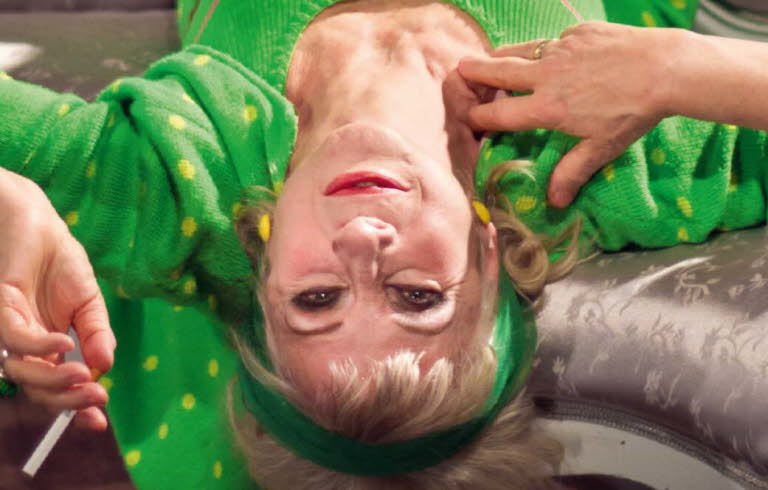Book cover of "Vem tror hon att hon är": it's a photo of a women lies upside down on a couch wearing a green sweatsuit with green dots, smoking a cigarette.