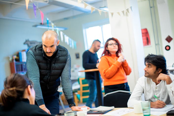 Fem vuxna rör sig i ett rum som påminner om ett klassrum. En man står upp och tittar på två sittande personer. Det ser ut att vara en kurs för vuxna.