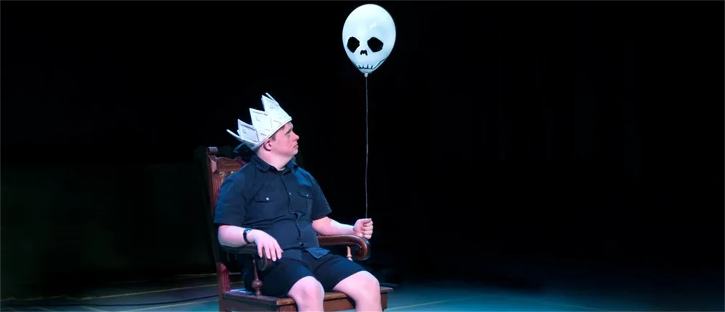 En man sitter på en scen. På huvudet har han en kungakrona i papp, i handen håller han en ballong med spökansikte.