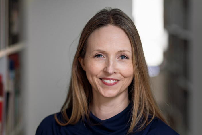 Medelålders kvinna med långt cendréfärgat hår och blå tröja omgiven av bokhyllor.