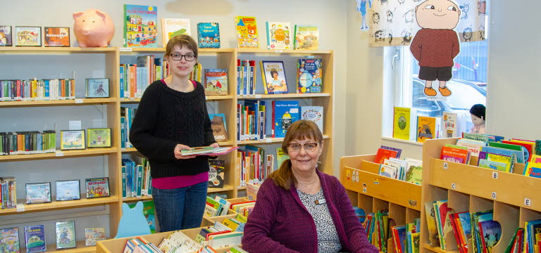 Två kvinnor, en sittande och en stående, står i bibliotekets barnboksområde. En bild av Alfons Åberg hänger vid fönstret.