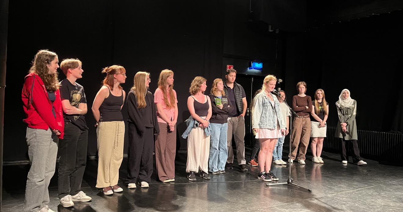 En rad ungdomar står på en scen, en ung kvinna står lite längre fram vid en mikrofon.