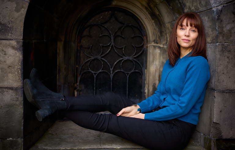 Lina Wolff sits in a window with her legs up, she is looking into the camera and wearing a blue shirt.