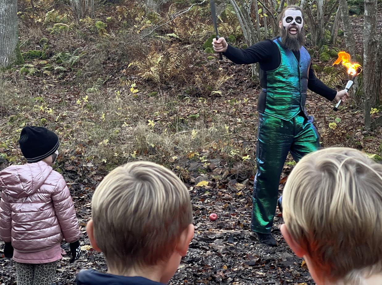 Barn som tittar på en artist i grönblank kostym med en fackla i handen ute i skogen. 