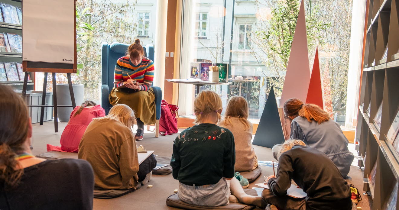 En grupp barn sitter utspridda framför en kvinna i en fåtölj, alla sitter böjda över sina skrivböcker.