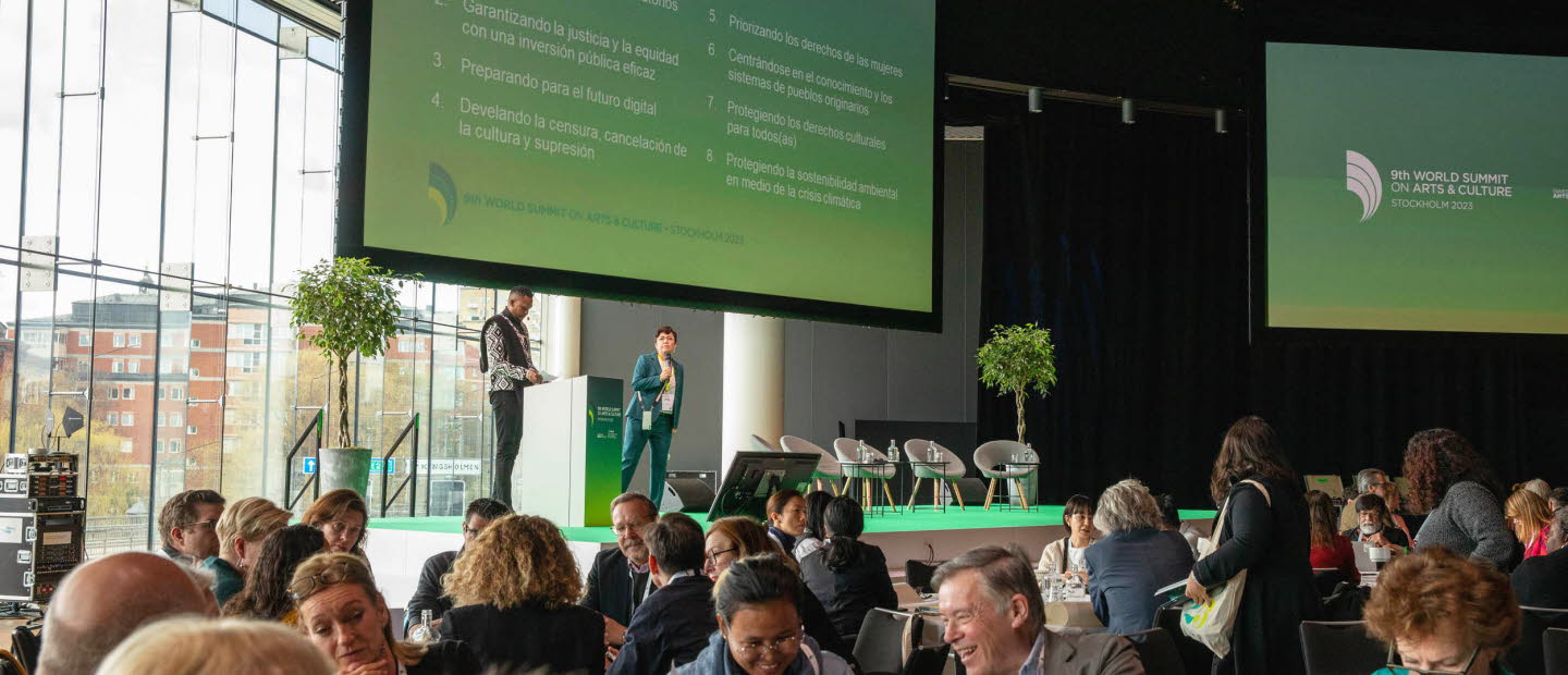 Vy över konferenslokalen och deltagare som sitter i grupper kring bord. På scenen två talare och en stor skärm som visar en presentation. 