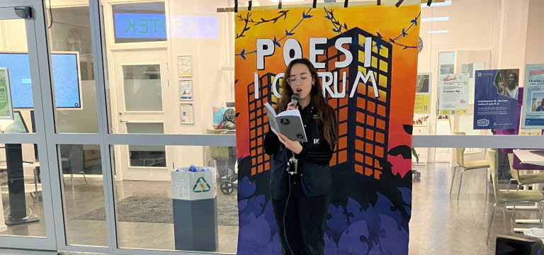 En ung kvinna står med en mikrofon i ett köpcentrum och läser ur en bok.