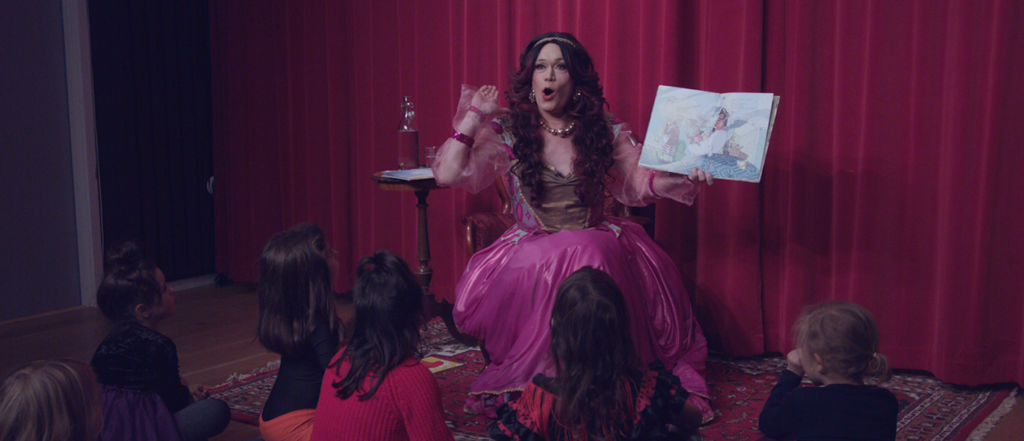 En dragqueen som läser högt för en grupp barn som sitter på golvet. 