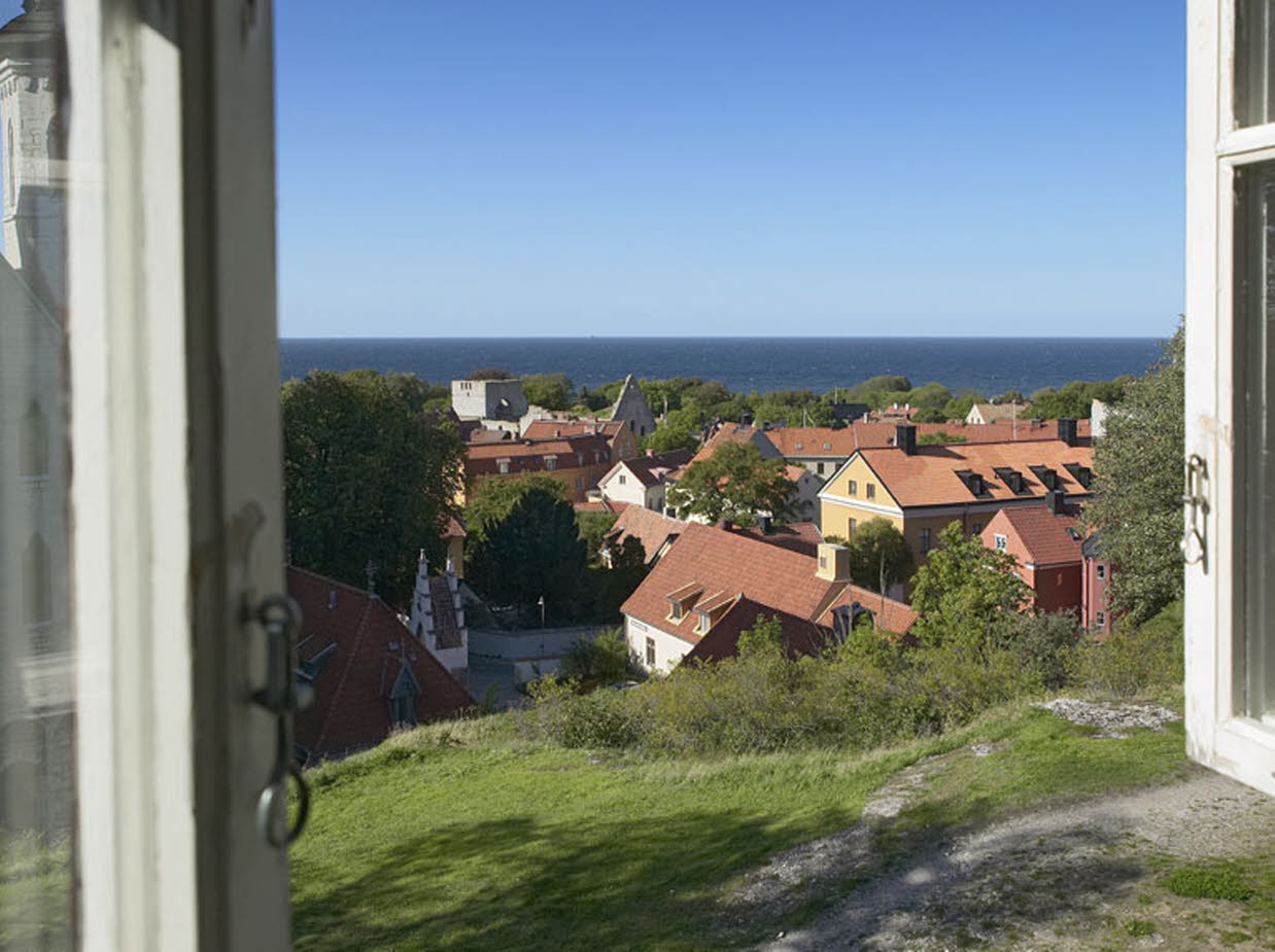 Ett öppet träfönster visar en vy över hustak, i horisonten syns havet som möter en blå himmel. Huset ligger högt och det syns lite berg och gräs precis utanför fönstret.